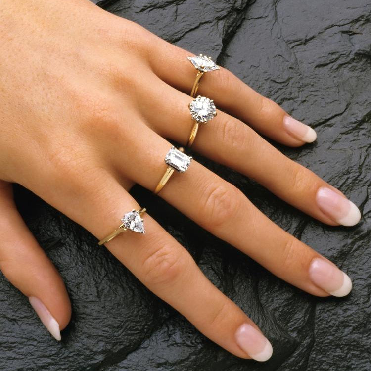 Four 14K gold diamond rings with emerald, round, and trillion cuts on a hand against dark stone